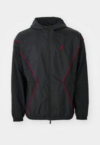 Sort hættejakke med lynlås foran, røde pipingdetaljer langs siderne og skuldrene, samt et lille rødt Air Jordan-logo på brystet.