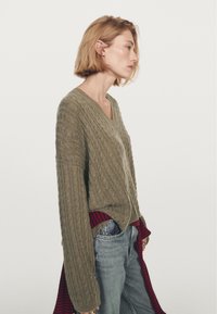 Olivgrön kabelstickad tröja med V-ringning och långa ärmar, lagd över blå jeans och accentuerad med en röd och marinblå rutig skjorta knuten i midjan.