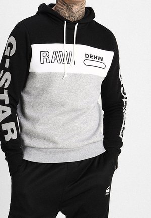 Homme portant un sweat à capuche noir, blanc et gris avec "RAW DENIM" sur la poitrine et "G-STAR" sur les manches, assorti d'un pantalon noir G-Star.