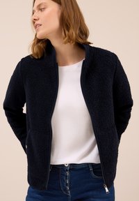 Granatowa polarowa kurtka z wysokim kołnierzem, zamek błyskawiczny i boczne kieszenie. Strukturalny materiał zapewnia ciepło i komfort. Widoczne białe top i dżinsy.