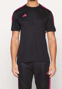 Svart atletisk t-shirt med korta ärmar, kontrasterande rosa ränder på axlarna och en liten rosa logotyp på vänster bröst. Slät textur.