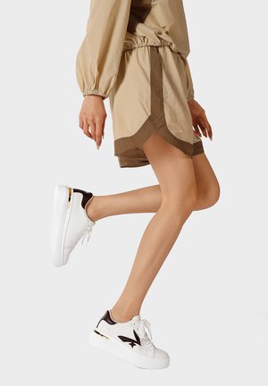 Gambe con pantaloncini beige e sneakers bianche con dettagli marroni, in posa a metà passo su uno sfondo chiaro e uniforme.