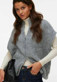 Kvinna med långt brunt hår som bär en grå stickad ärmlös zip-up tröja över en krämfärgad polotröja, håller en svart väska och poserar med handen i fickan.