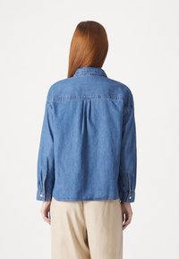 Personne aux longs cheveux roux porte une chemise en jean bleue et un pantalon beige, vue de dos sur un fond blanc uni.