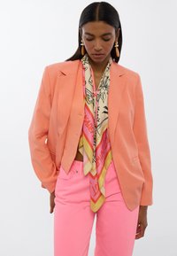 Korallenblazer mit strukturierter Passform, leichtem Stoff; kombiniert mit einem gemusterten Seidenschal in Rosa, Gelb und Creme. Rosa Hosen vervollständigen den Look.