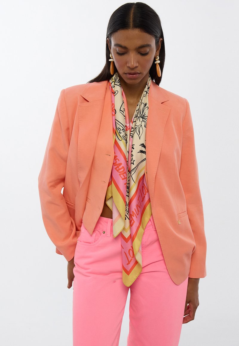 Korallenblazer mit strukturierter Passform, leichtem Stoff; kombiniert mit einem gemusterten Seidenschal in Rosa, Gelb und Creme. Rosa Hosen vervollständigen den Look.