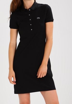 Frau trägt ein enganliegendes, schwarzes Polo-Kleid mit kurzen Ärmeln, Kragen und Knöpfen, mit einem kleinen weißen Logo auf der Brust.