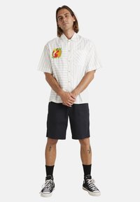 Korte mouwen witte shirt met dunne zwarte strepen, voorzien van een kleurrijke fruitafbeelding op de linkerkant van de borst. Gecombineerd met donkere shorts en sneakers.