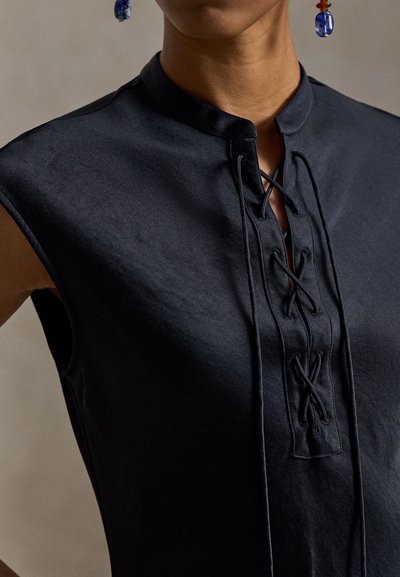 Μαύρο τοπ με κοντά μανίκια, ψηλό γιακά και κορδόνια μπροστά. Λεία υφή με διακριτική λάμψη, διακοσμημένο με σταυρωτές λεπτομέρειες από ύφασμα.