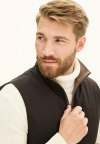 Schwarze Reißverschlussweste mit einem braunen Kragen, getragen über einem weißen gerippten Rollkragenpullover. Glatte Stoffqualität mit taillierter Passform, die einen praktischen Look präsentiert.