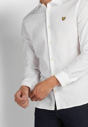 Mand iført hvid skjorte med knapper og gul fuglelogo på brystet, som justerer manchet, kombineret med mørke bukser.