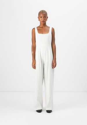 Une combinaison en lin blanc avec un décolleté carré, une silhouette à jambes larges et des coutures minimales. Le modèle se tient droit, portant des chaussures noires et des boucles d'oreilles en forme de créole.