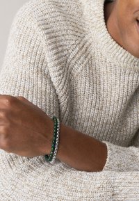 Bracciale di perline con pietre verdi e accenti in metallo argentato, indossato su un polso accanto a un pullover beige a maglia testurizzata.