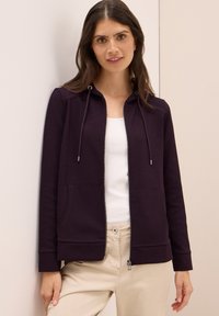 Felpa con zip di colore viola scuro, con cappuccio regolabile, due tasche frontali e polsini a costine, abbinata a una canottiera bianca e pantaloni beige.