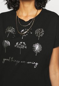 Musta t-paita, jossa hopeanvärisiä voikukan kuvia ja teksti "good things are coming" kaunokirjoitusfontilla. Lyhyet hihat, rento istuvuus.