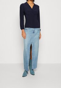 Marinblå omslagsblus med långa ärmar, i kombination med en ljusblå denimmaxikjol med en framklippning och fransig fåll. Matchas med turkosa ankelboots.