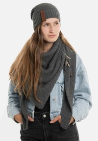 Grijze gebreide beanie en een oversized sjaal in shawl-stijl, beide getextureerd, gedragen met een spijkerjack, met een decoratieve speldaccent.