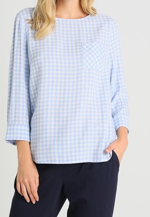 Frau trägt eine hellblau-weiß karierte Bluse mit langen Ärmeln und einer kleinen Brusttasche sowie eine dunkelblaue Hose.