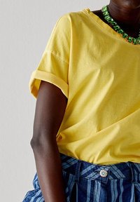 T-shirt di cotone giallo con maniche corte arrotolate, abbinato a pantaloni corti a vita alta blu e bianchi a righe con un bottone decorativo.
