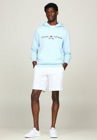 Sudadera azul claro con cordones blancos y logo, combinada con pantalones cortos blancos y zapatillas blancas. Tejido suave, corte relajado, diseño casual.