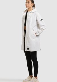 Donna che indossa un impermeabile bianco con cappuccio sopra leggings neri e un top corto nero, in piedi con una mano in tasca.