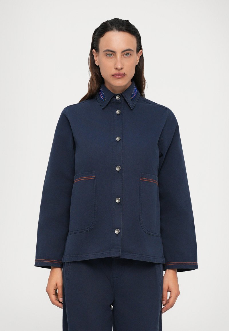 Άτομο που φοράει ένα ναυτικό μπλε σακάκι με κουμπιά, το οποίο έχει μπροστινές τσέπες και κεντημένο γιακά, στέκεται μπροστά από έναν απλό λευκό φόντο.