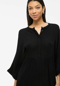 Robe noire en tissu texturé avec un col henley, des manches trois-quarts, et une coupe ample avec un design plissé.