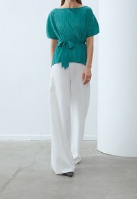 Teal plisserad blus med korta ärmar och knytbart midjeband, matchad med vita vida byxor; textil med struktur och flödande design.