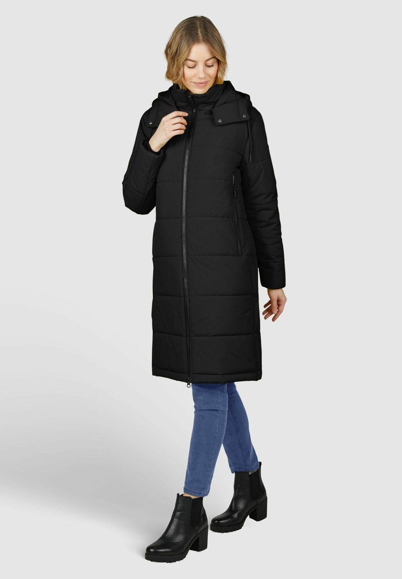 Schwarze gepolsterte Jacke mit Kapuze, Reißverschluss und seitlichen Taschen. Sie hat einen langen, geraden Schnitt und wird mit blauen Jeans und schwarzen Stiefeletten kombiniert.