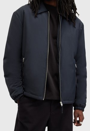 Person iført en mørk marineblå lynlåstrøje med elastiske manchetter og lynlåslommer i siden, kombineret med sorte bukser og en mørk skjorte.