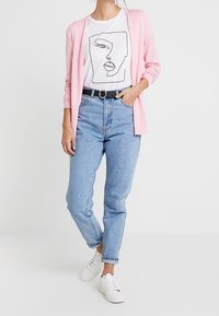 Cardigan rose porté sur un t-shirt blanc avec un dessin au trait noir, associé à un jean bleu taille haute et des baskets blanches.