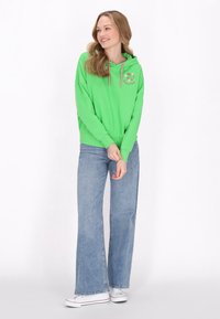 Felpa verde con cordini rosa, con ricamo floreale sul petto, abbinata a jeans a gamba larga blu e sneakers bianche.