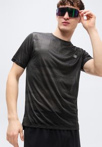 Μαύρο αθλητικό πουκάμισο με κοντά μανίκια, διαθέτει υφασμάτινο μοτίβο, στενή εφαρμογή και μικρό λογότυπο στο στήθος. Φορεμένο με μεγάλα ανακλαστικά γυαλιά ηλίου.