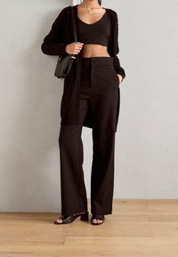 Top corto negro sin mangas, cárdigan largo negro y pantalones negros de pierna ancha. Tejido liso con un ajuste relajado y hardware mínimo. Sandalias marrones de tacón.