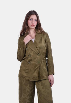 Giovane donna che indossa un blazer doppiopetto a fantasia floreale verde oliva e pantaloni ampi coordinati, in piedi davanti a uno sfondo semplice.