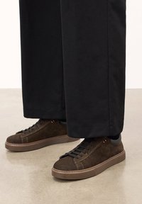 Zapatillas de ante marrón con detalles en negro, diseño plano con cordones y suela de goma texturizada, combinadas con pantalones negros de pierna ancha.