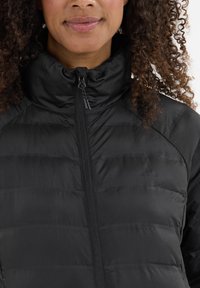Sort pufferjakke med høj krave, der har en lynlås foran og et quiltet design med vandrette syninger. Blødt, letvægtsmateriale.