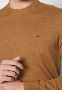 Brun stickad tröja med rundad halsringning, ribbade ärmslut, med en liten logotyp i blått, vitt och rött på bröstet.