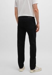 Hombre con jeans rectos negros, camiseta blanca y zapatillas blancas de pie contra un fondo gris claro liso.