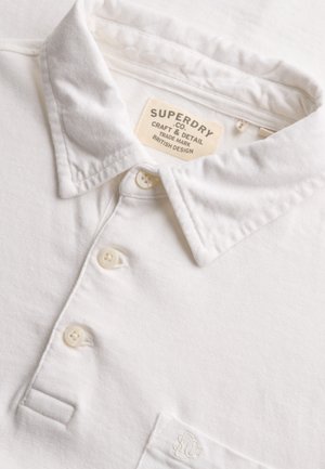 Polo bianca con collo a costine e tre bottoni, etichetta del marchio Superdry cucita all'interno del collo e ricamo discreto del logo sulla tasca del petto.
