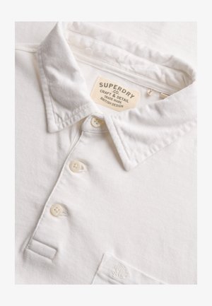 Polo bianca con collo a costine e tre bottoni, etichetta del marchio Superdry cucita all'interno del collo e ricamo discreto del logo sulla tasca del petto.