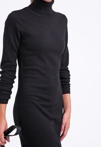 Svart polokrageklänning med långa ärmar, figurnära design, slät textur, når till mitten av vad; hålls med en cirkulär handtagsaccessoar.