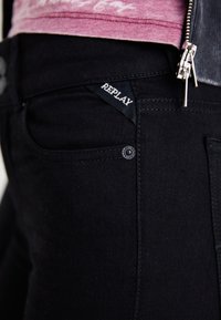 Black denim jeans with a tonal stitch. Features a small "REPLAY" label on the waistband and a metal button at the front pocket.