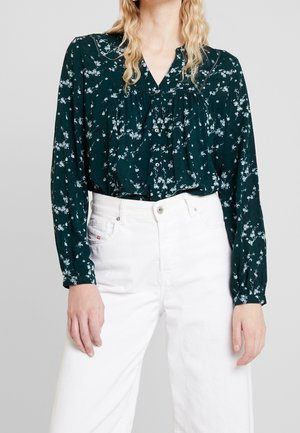 Femme portant un chemisier à manches longues à fleurs vert foncé, rentré dans un jean blanc taille haute, visible des épaules aux hanches.