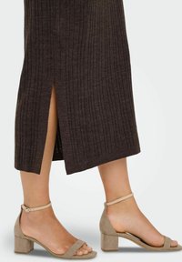 Underben i beige ankelrem blokkede hæle sandaler og en mørk ribbet nederdel med en slids bagpå mod en lys baggrund.
