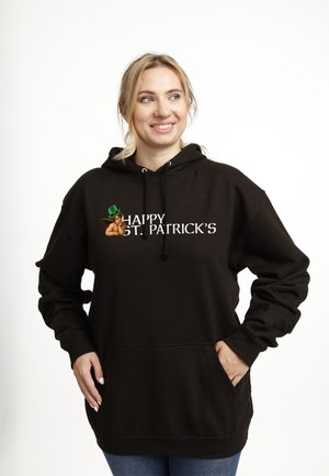 Smilende kvinde har en sort hættetrøje på med teksten "Happy St. Patrick’s" og en kerub-emoji med grøn hat, stående foran en ensfarvet baggrund.
