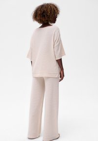 Vrouw met krullend haar, gekleed in een losse, crèmekleurige gebreide top en wijde broek, staande tegen een effen witte achtergrond en zijwaarts gekeerd.