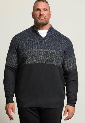 Homme barbu portant un pull à col zippé bleu foncé et noir, debout devant un fond clair uni.