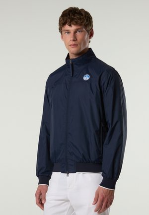 Hombre con chaqueta con cremallera azul marino y un logo circular en el pecho, combinado con pantalones blancos, posando frente a un fondo neutro.