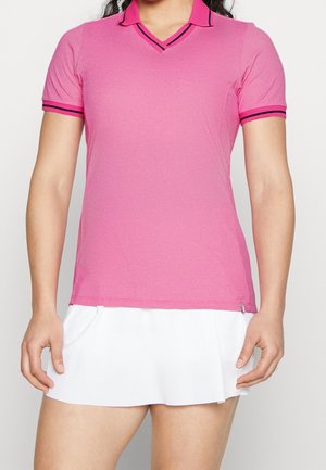 Kvinde iført en pink polo-shirt med korte ærmer med mørk kant og en hvid tennisnederdel, stående mod en ensfarvet lys baggrund.